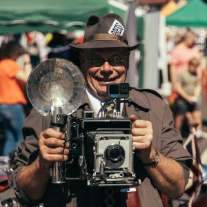A man wearing 1940's suit with a 1940's hat with a press card in the band is holding a war-time camera looking at the person taking a photo
